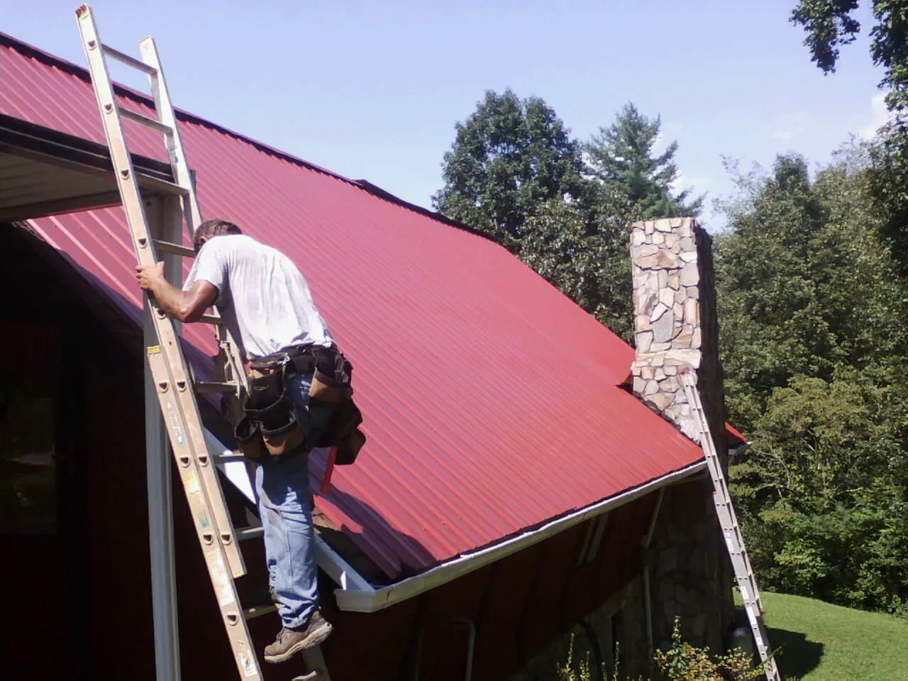 Roofer on a ladder beside a red metal roof and stone chimney — Soffit & Fascia Repair by Roofing Brothers in Sibley